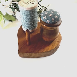 May include: A heart-shaped wooden base holds a spool of blue-grey thread and a small wooden container topped with a dark teal pincushion featuring a repeating pattern of tiny sewing spools. A small wooden peg stands upright on the base.