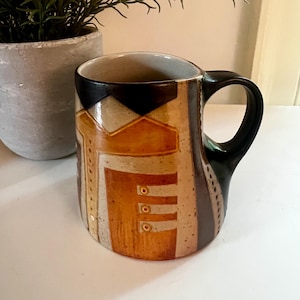 May include: A ceramic mug with a black handle and rim, featuring a geometric design in shades of brown, beige, and orange. The mug has a cylindrical shape and is placed on a white surface.