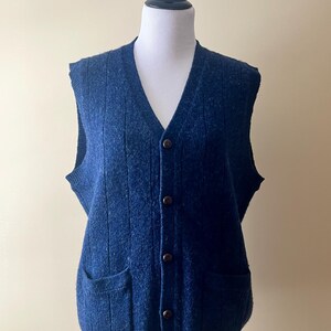 May include: A navy blue, button-down sweater vest with a V-neck and two front pockets. The vest is made of a textured knit fabric and features brown buttons. The garment is displayed on a white mannequin.