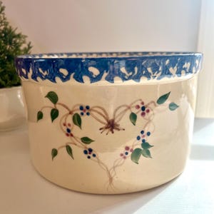 May include: Cream-colored ceramic bowl with a blue scalloped rim. The bowl is decorated with a floral design featuring green leaves, blue and red berries, and a brown bow.