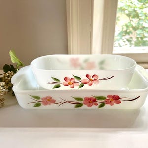 May include: Two white, rectangular casserole dishes with rounded corners. Each dish is decorated with pink flowers and green leaves. The dishes are stacked on top of each other, with the smaller dish on top.