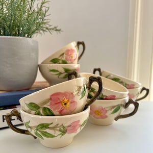 May include: A set of six cream-colored teacups with a floral design. Each cup features a pink flower with yellow center and green leaves. The handles are a dark brown color. The cups are stacked on a white surface.