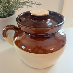 May include: A two-tone brown and cream-colored ceramic crock with a lid and handle. The lid has a small, round, light-colored wooden knob. The crock is likely used for food storage or as a decorative kitchen item.