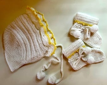 May include: A handmade, off-white crochet baby bonnet and matching booties set. The bonnet has a scalloped edge with yellow trim, and the booties feature yellow accents and white pom-poms. A charming, cozy ensemble.
