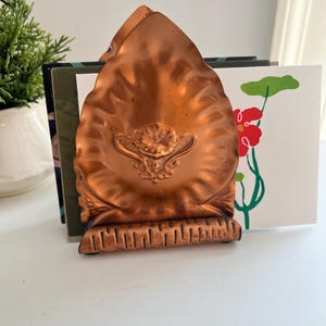 May include: A copper-colored bookend with an ornate, embossed design. The bookend has a fan-like shape with a decorative base. It is holding several books and a card with a red flower illustration.