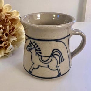 May include: A beige ceramic mug with a dark blue horse illustration. The mug has a rounded shape with a handle and a dark blue band around the top and bottom of the horse design. The mug is on a white surface.