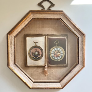 May include: An octagonal wall frame with a distressed gold and white finish, featuring a top metal hanger. Inside, a book-shaped display shows two circular pocket watch illustrations on a beige fabric background, accented by a small hanging tassel.