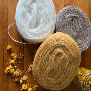May include: Three skeins of yarn in white, brown, and golden yellow sit on a wooden surface. Dried yellow flowers and a glass jar filled with flowers are scattered around the yarn. The yarn is tightly wound, showing the texture of the fibers.