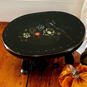 May include: An oval black footstool with a glossy finish, featuring a central inlay design of a basket filled with flowers and a bird. The border is decorated with a repeating diamond pattern, and the stool stands on four short, dark wooden legs.