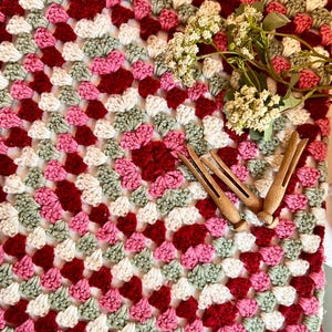 May include: A handmade crochet blanket in a granny square pattern. The blanket features squares in shades of white, pink, red, and green. Wooden clothespins and sprigs of white flowers are placed on top of the blanket.