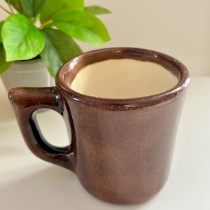 May include: A brown ceramic mug with a rounded handle. The mug has a dark brown exterior and a cream-colored interior. The mug is sitting on a white surface, with a green plant in the background.
