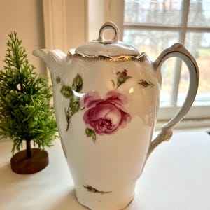 May include: White porcelain teapot with a lid, featuring a large pink rose and smaller rose buds. The handle and rim have gold accents. A small decorative evergreen tree is in the background.