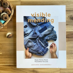 May include: A book titled "Visible Mending" with a cover featuring a close-up of patched denim jeans. The book's subtitle reads "Repair, Renew, Reuse The Clothes You Love" by Arounna Khounnoraj. The book is on a wooden surface.