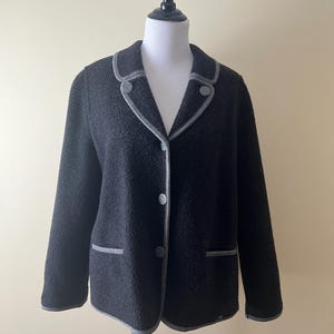 May include: A black jacket with a textured fabric, featuring a gray trim along the collar, lapels, pockets, and cuffs. The jacket has three gray buttons down the front and is displayed on a mannequin.