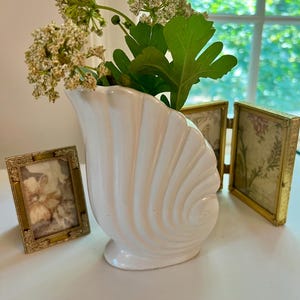 May include: A white seashell-shaped vase holds greenery and flowers. Two small, gold-framed pictures are on the left and right. The vase is on a white surface, with a window in the background.