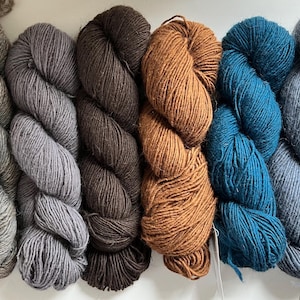 May include: A collection of eight skeins of yarn in various colors, including brown, gray, orange, teal, and blue. The yarn is tightly wound and appears soft, suitable for knitting or crocheting projects. The skeins are arranged side by side.