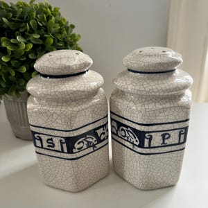 May include: A pair of ceramic salt and pepper shakers with a crackle glaze finish. The shakers are off-white with dark blue accents and lettering. The lids have multiple holes for dispensing.