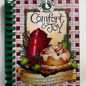 May include: A spiral-bound cookbook titled Comfort & Joy by Gooseberry Patch. The cover features a red coffee pot, a cinnamon roll, and holly on a red and white plaid background. Text reads: Cozy Christmas recipes, favorite holiday memories.