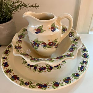 May include: A cream-colored ceramic pitcher and a matching wide, shallow bowl with a ruffled edge. Both pieces feature a hand-painted pattern of dark blue berries, green leaves, and small white flowers arranged in a continuous vine motif.