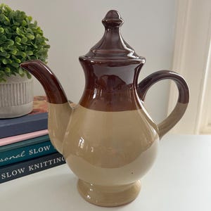 May include: A ceramic teapot with a brown and tan color scheme. The teapot has a bulbous body, a curved spout, and a handle. The lid is topped with a decorative finial. The teapot is sitting on a white surface.