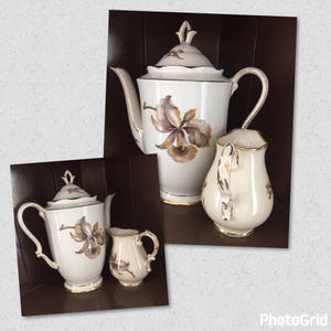 May include: Cream-colored porcelain tea set with a floral design in shades of brown and gold. The set includes a teapot, creamer, and sugar bowl, all with gold trim. The teapot has a lid and a curved spout.