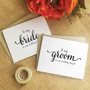 Puede incluir: Dos tarjetas blancas con caligraf&iacute;a negra que dice "To my bride on our wedding day" y "To my groom on our wedding day". Cada tarjeta tiene un peque&ntilde;o coraz&oacute;n rojo.