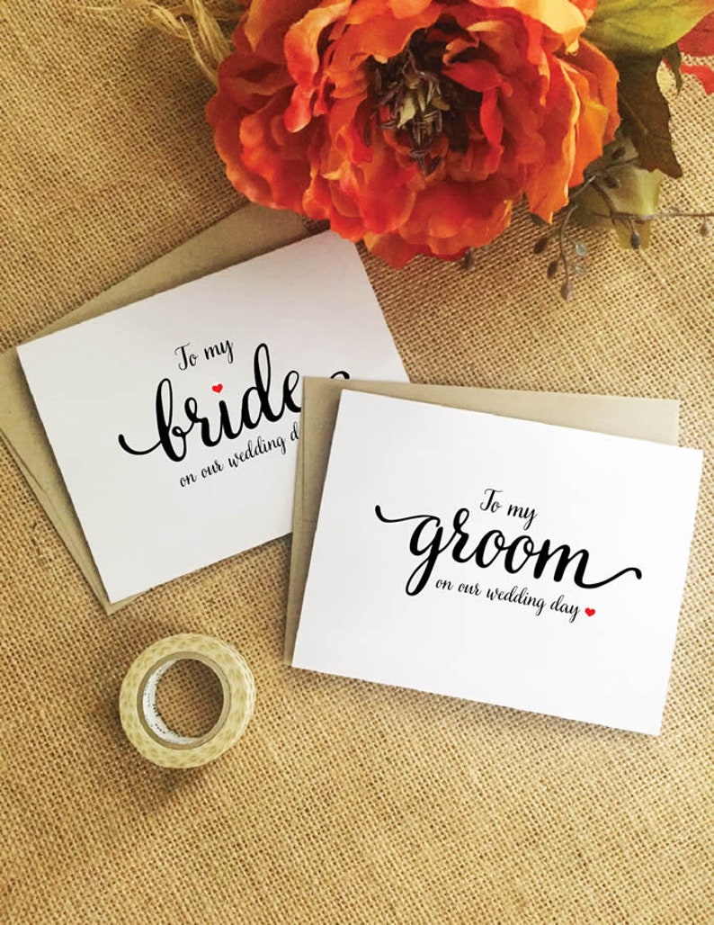 Puede incluir: Dos tarjetas blancas con caligraf&iacute;a negra que dice "To my bride on our wedding day" y "To my groom on our wedding day". Cada tarjeta tiene un peque&ntilde;o coraz&oacute;n rojo.