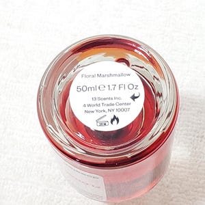 Puede incluir: Una botella de vidrio transparente con l&iacute;quido rojo en su interior. La etiqueta dice "Floral Marshmallow" y "50ml e 1.7 Fl Oz." La botella es de 13 Scents Inc., ubicada en Nueva York, NY. La parte inferior de la botella tiene un s&iacute;mbolo de advertencia.