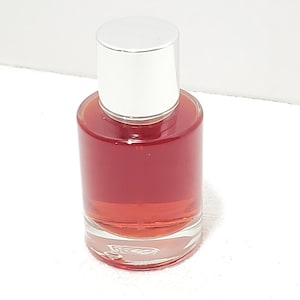 Puede incluir: Un frasco de perfume de vidrio transparente con tapa plateada. La botella est&aacute; llena de un l&iacute;quido rojizo-rosado. La botella tiene forma cil&iacute;ndrica y se apoya sobre una superficie blanca. La tapa es de color plateado brillante.
