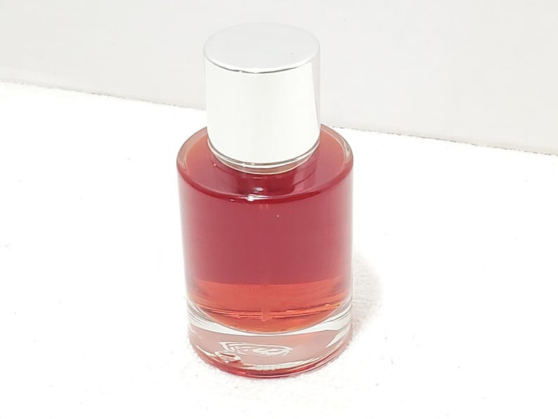 Puede incluir: Un frasco de perfume de vidrio transparente con tapa plateada. La botella est&aacute; llena de un l&iacute;quido rojizo-rosado. La botella tiene forma cil&iacute;ndrica y se apoya sobre una superficie blanca. La tapa es de color plateado brillante.