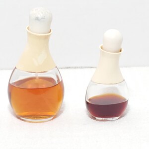 May include: Two glass perfume bottles with beige tops and clear, rounded bases. One bottle contains amber liquid, the other a dark red liquid. Both have white stoppers and sit on a white surface.