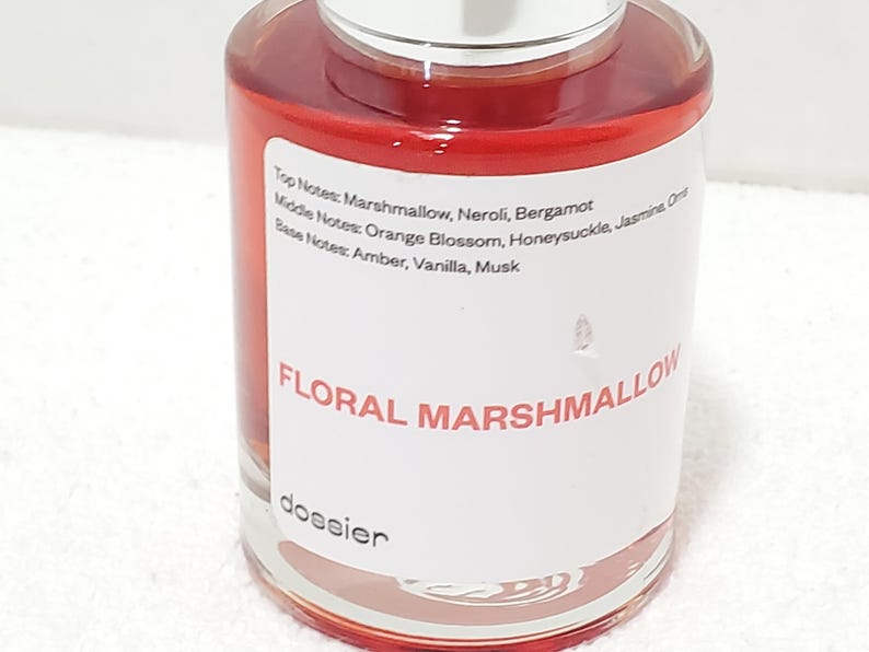 Puede incluir: Un frasco de perfume transparente con tapa plateada, lleno de un l&iacute;quido rojo-anaranjado. La etiqueta dice "FLORAL MARSHMALLOW" y enumera las notas de fragancia. La marca "dossier" tambi&eacute;n es visible.