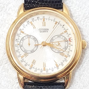 May include: A gold-tone Citizen Quartz watch with a round cream-colored dial, Roman numeral hour markers, and two sub-dials displaying days of the week and months. The watch features a date ring around the perimeter and a black textured leather strap.