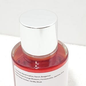 Puede incluir: Un frasco de perfume con tapa plateada y cuerpo de vidrio transparente. El l&iacute;quido interior es de color rojo anaranjado. La etiqueta enumera las notas de salida, coraz&oacute;n y fondo de la fragancia.