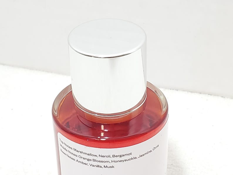 Puede incluir: Un frasco de perfume con tapa plateada y cuerpo de vidrio transparente. El l&iacute;quido interior es de color rojo anaranjado. La etiqueta enumera las notas de salida, coraz&oacute;n y fondo de la fragancia.