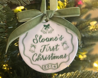 Può includere: Una decorazione natalizia in ceramica bianca con testo e dettagli verdi, con la scritta "Sloane's First Christmas" e l'anno "2024". Adornata con un nastro e un fiocco di velluto verde, appesa a un albero di Natale.