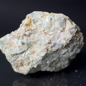 May include: A close-up of a rough, irregularly shaped rock. The rock has a light beige and off-white base color with patches of pale blue and tan. The surface appears textured and granular, with visible crystalline structures.