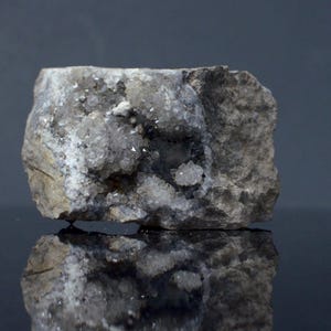 May include: A raw, jagged rock specimen featuring a central cavity filled with small, sparkling, translucent crystalline formations. The exterior of the stone is textured, matte, and greyish-brown, contrasting with the bright, reflective mineral clusters.