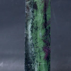 May include: A polished stone tower with a pointed top, featuring a vertical composition of vibrant green, deep black, and dark magenta mineral inclusions. The surface displays natural crystalline textures and varied color bands throughout the structure.