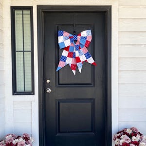 May include: A large decorative bow made of fabric squares in a patchwork pattern of red, white, and blue. The bow features various prints including stars, small dots, and geometric grid patterns, hanging centered on a solid black front door.