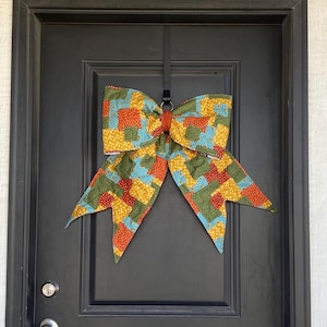 May include: A large fabric bow featuring a patchwork pattern of squares in shades of orange, yellow, green, and light blue with small white polka dots. The bow is secured with a solid orange center band and hangs from a black metal door hook.