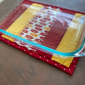 May include: A glass baking dish with a light blue rim sits on a red and yellow quilted hot pad with white polka dots.