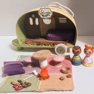May include: A miniature wooden camper with a welcome sign, two wooden dolls, a purple cushion, a table with two mushroom stools, a fire pit, and a picnic blanket with cookies and marshmallows.