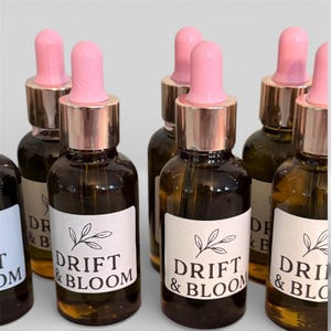 May include: A collection of small, dark amber glass bottles with pink dropper tops and gold-colored collars. Each bottle has a white label with the text "DRIFT & BLOOM" and a leaf design. The bottles are arranged closely together.