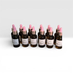 May include: A collection of small, dark glass bottles with pink droppers and gold-colored collars. Each bottle has a white label with the words "DRIFT & BLOOM" and a leaf design. The bottles are arranged on a white surface.