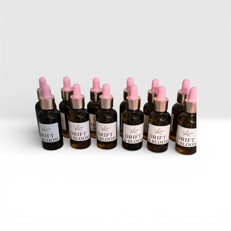 May include: A collection of small, dark glass bottles with pink droppers and gold-colored collars. Each bottle has a white label with the words "DRIFT & BLOOM" and a leaf design. The bottles are arranged on a white surface.