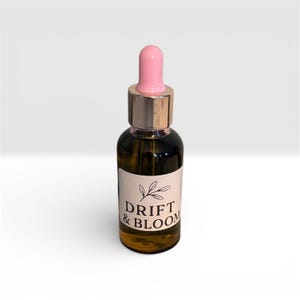 May include: A small, dark glass bottle with a pink dropper and gold-colored cap. The bottle has a white label with the words "DRIFT & BLOOM" and a leaf design. The bottle is filled with a golden liquid.