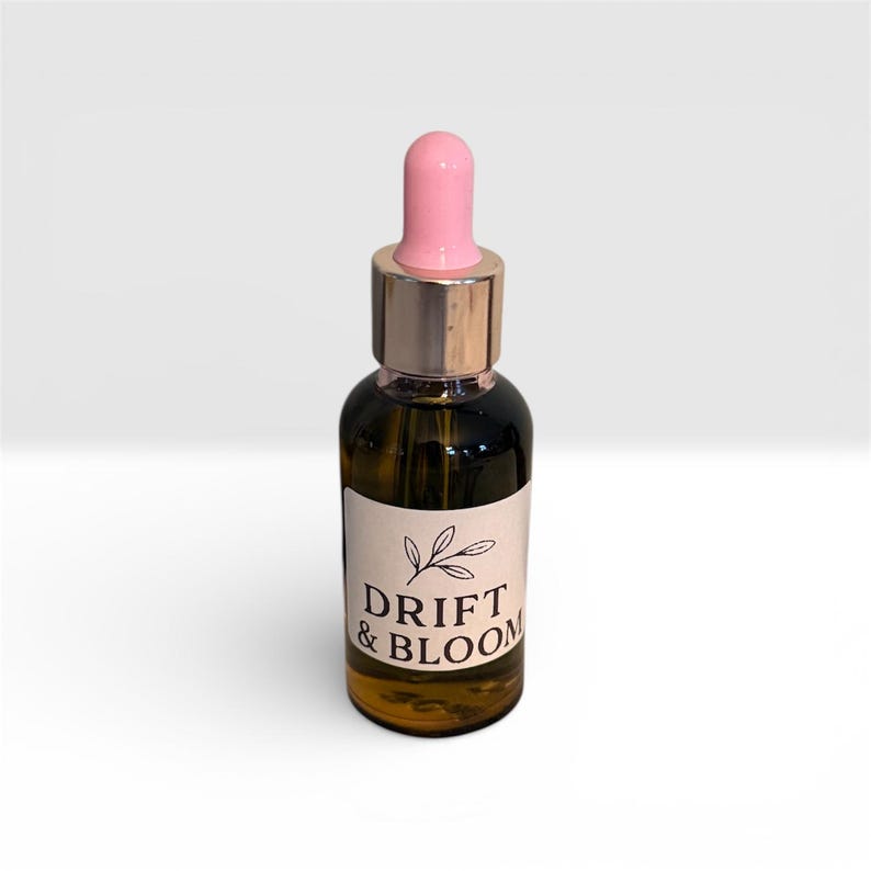 May include: A small, dark glass bottle with a pink dropper and gold-colored cap. The bottle has a white label with the words "DRIFT & BLOOM" and a leaf design. The bottle is filled with a golden liquid.
