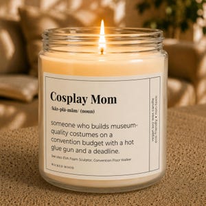 May include: A clear glass jar candle with a lit wick and a white label featuring the text Cosplay Mom, a phonetic pronunciation, a definition, and the brand name Wicked Mood. The label also lists vegan soy wax, eco-friendly, and non-toxic.