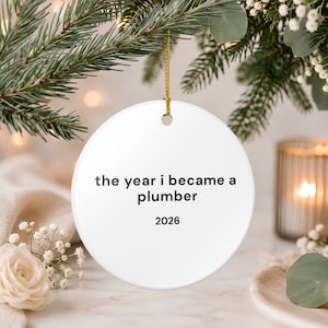 May include: A round white ceramic ornament hanging from a gold string. The ornament features black text that reads "the year i became a plumber" followed by the year "2026" centered below the text on the smooth surface of the circular object.